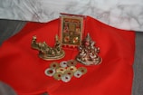A red fabric surface is adorned with religious artifacts, including two small metallic statues and a decorative plaque. Several coin-shaped objects with a golden hue are scattered around.
