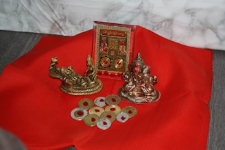 A red fabric surface is adorned with religious artifacts, including two small metallic statues and a decorative plaque. Several coin-shaped objects with a golden hue are scattered around.
