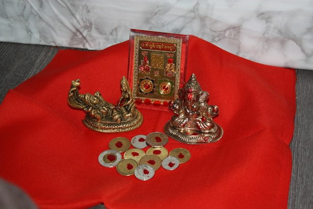 A red fabric surface is adorned with religious artifacts, including two small metallic statues and a decorative plaque. Several coin-shaped objects with a golden hue are scattered around.