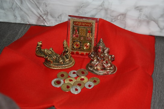 A red fabric surface is adorned with religious artifacts, including two small metallic statues and a decorative plaque. Several coin-shaped objects with a golden hue are scattered around.