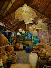 A cozy shop interior with earthy tones, featuring a variety of large clay pots arranged on the floor. Overhead, there are numerous woven pendant lights creating a warm, inviting glow. The shop's structure suggests a rustic, natural aesthetic with wooden beams and a thatched roof. The area is filled with objects including small potted plants, a blue plastic stool, and a backdrop of a brick-patterned wall.