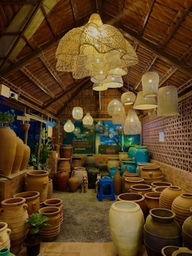 A cozy shop interior with earthy tones, featuring a variety of large clay pots arranged on the floor. Overhead, there are numerous woven pendant lights creating a warm, inviting glow. The shop's structure suggests a rustic, natural aesthetic with wooden beams and a thatched roof. The area is filled with objects including small potted plants, a blue plastic stool, and a backdrop of a brick-patterned wall.