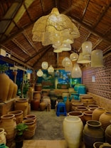A cozy shop interior with earthy tones, featuring a variety of large clay pots arranged on the floor. Overhead, there are numerous woven pendant lights creating a warm, inviting glow. The shop's structure suggests a rustic, natural aesthetic with wooden beams and a thatched roof. The area is filled with objects including small potted plants, a blue plastic stool, and a backdrop of a brick-patterned wall.