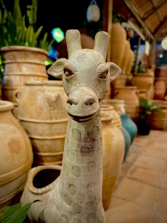 A ceramic sculpture of a giraffe with detailed facial features. The background consists of large terracotta pots and some greenery, creating an earthy, natural setting.
