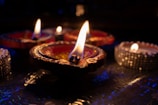 A close-up view of several decorative oil lamps with flames flickering gently in a dimly lit setting, creating a warm and intimate ambiance. The lamps are adorned with intricate designs and vivid colors, set against a subtly reflective surface.