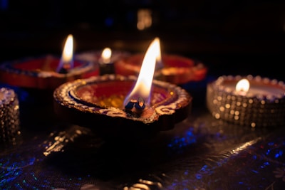 A close-up view of several decorative oil lamps with flames flickering gently in a dimly lit setting, creating a warm and intimate ambiance. The lamps are adorned with intricate designs and vivid colors, set against a subtly reflective surface.