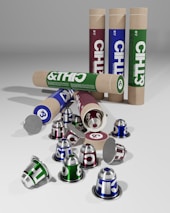 Several cylindrical cardboard tubes and coffee capsules in green, blue, and maroon are seen. The tubes are standing upright and lying on their side, with some capsules scattered around. The capsules are metallic and have bold lettering.