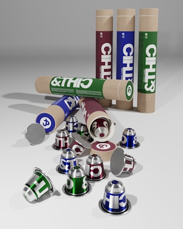 Several cylindrical cardboard tubes and coffee capsules in green, blue, and maroon are seen. The tubes are standing upright and lying on their side, with some capsules scattered around. The capsules are metallic and have bold lettering.