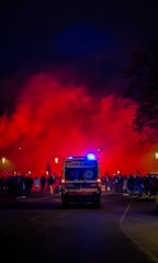 An ambulance with flashing blue lights is parked on a dark street. In the background, a large crowd of people is gathered, and red smoke or fog fills the air, casting an intense glow.