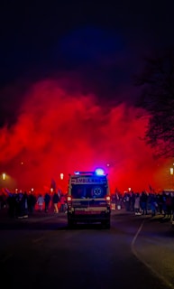 An ambulance with flashing blue lights is parked on a dark street. In the background, a large crowd of people is gathered, and red smoke or fog fills the air, casting an intense glow.