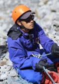 A person wearing an orange helmet, sunglasses, and a blue jacket is sitting on rocky terrain. They are holding climbing ropes and wearing gloves, appearing to be engaged in a mountaineering or outdoor climbing activity.