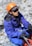 A person wearing an orange helmet, sunglasses, and a blue jacket is sitting on rocky terrain. They are holding climbing ropes and wearing gloves, appearing to be engaged in a mountaineering or outdoor climbing activity.