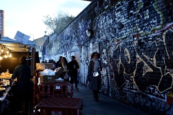 A bustling street market scene with a vendor stall on the left offering food, highlighted by warm overhead lights. Several people walk along a narrow pathway, bordered by a long brick wall covered in colorful graffiti art. The atmosphere is lively and urban, situated under an overcast sky with some visible greenery above the wall.