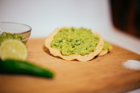 A close-up of a wooden board features a crunchy tortilla chip topped with a generous portion of mashed avocado, suggestive of guacamole. To the left, there is a partial view of a lime, while a green chili pepper and a small mound of salt are also nearby.
