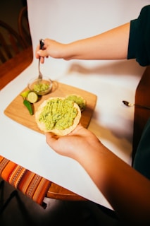 A hand holding a tostada topped with mashed avocado. A small jar and a cutting board with lime slices, jalapeño, and more avocado spread are visible in the background. The scene is set on a wooden table with a colorful cloth.