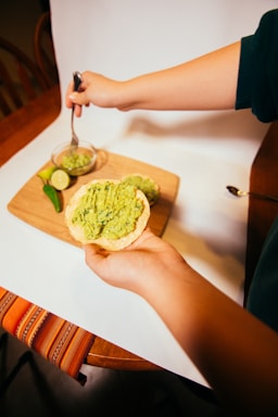 A hand holding a tostada topped with mashed avocado. A small jar and a cutting board with lime slices, jalapeño, and more avocado spread are visible in the background. The scene is set on a wooden table with a colorful cloth.