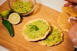 A wooden board displays a variety of ingredients for a Mexican dish. A glass bowl contains guacamole, while several tostadas are covered with guacamole and garnished with cilantro. Nearby, a jalapeño and a halved lime add color and flavor. A hand is holding a couple of plain tostadas.
