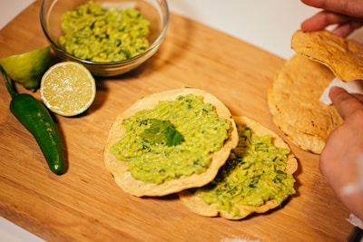 A wooden board displays a variety of ingredients for a Mexican dish. A glass bowl contains guacamole, while several tostadas are covered with guacamole and garnished with cilantro. Nearby, a jalapeño and a halved lime add color and flavor. A hand is holding a couple of plain tostadas.