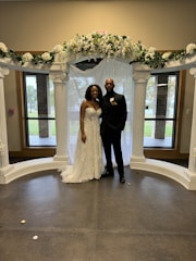 A couple is standing under a decorative arch adorned with white flowers and greenery. The woman is wearing an elegant white wedding dress with floral details, and the man is dressed in a black suit with a boutonniere. They are posing indoors with windows in the background showing an outdoor view.