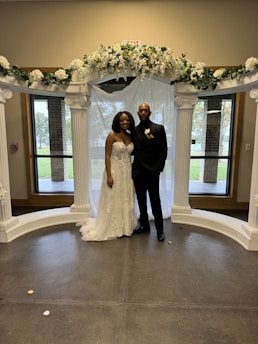 A couple is standing under a decorative arch adorned with white flowers and greenery. The woman is wearing an elegant white wedding dress with floral details, and the man is dressed in a black suit with a boutonniere. They are posing indoors with windows in the background showing an outdoor view.