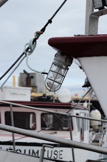 A close-up view of a boat's exterior, featuring a glass-encased light fixture attached to the side. Ropes and metallic structures are seen in the foreground, with parts of another boat visible in the background. The sky is overcast, contributing to a muted color palette.