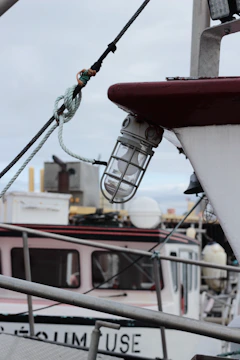 A close-up view of a boat's exterior, featuring a glass-encased light fixture attached to the side. Ropes and metallic structures are seen in the foreground, with parts of another boat visible in the background. The sky is overcast, contributing to a muted color palette.