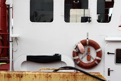 The image depicts the side of a ship with a white and red color scheme. A life preserver ring is attached to the side, with a nearby small, rectangular window. The life preserver has lettering on it and is accompanied by other nautical equipment such as coiled rope and fenders, against the backdrop of a metal structure.