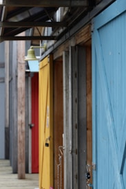 A series of colorful wooden doors, including blue, yellow, and red, line a narrow passageway. The doors are slightly open and show hinges and locks. Above them, a metal structure with beams is visible. A distinct yellow outdoor lamp hangs prominently above the yellow door.