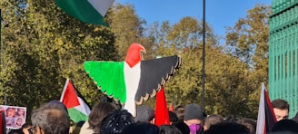 A large crowd of people gathered outdoors, many holding flags with red, white, green, and black colors. A wooden sculpture shaped like a bird with similar colors is prominently displayed above the crowd. The background features green foliage under a clear blue sky.