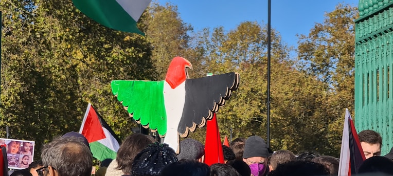 A large crowd of people gathered outdoors, many holding flags with red, white, green, and black colors. A wooden sculpture shaped like a bird with similar colors is prominently displayed above the crowd. The background features green foliage under a clear blue sky.
