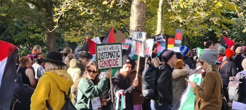 A group of people participating in a protest are shown holding various signs and flags. The signs include messages related to systemic genocide, Palestine, and Free Palestine. The crowd is gathered in an outdoor area surrounded by trees with sunlight filtering through leaves. Most people are wearing jackets and hats, indicating cooler weather.