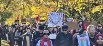 A large group of people marching through a park, carrying various signs and flags. Many signs include slogans and messages related to armistice and freedom, and several Palestinian flags are visible. The background is filled with trees displaying autumn foliage.
