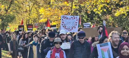 A large group of people marching through a park, carrying various signs and flags. Many signs include slogans and messages related to armistice and freedom, and several Palestinian flags are visible. The background is filled with trees displaying autumn foliage.