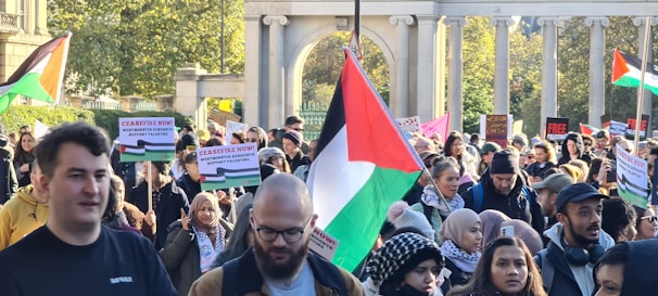 A large group of people gathered for a protest, holding signs and flags. The flags are red, green, black, and white, and the signs have messages related to ceasefire and support for Palestine. The gathering is set outdoors, with trees and a stone colonnade in the background.