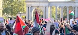 A large group of people are participating in a protest or demonstration. Many individuals are holding flags and signs with various messages. The scene is outdoors, and the background includes trees and a structure with columns.