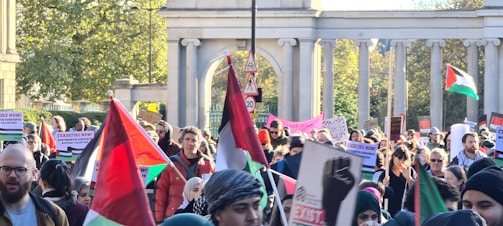 A large group of people are participating in a protest or demonstration. Many individuals are holding flags and signs with various messages. The scene is outdoors, and the background includes trees and a structure with columns.