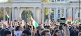 A large crowd of people gathered in a public space, many holding flags and signs advocating for Palestine. The scene is framed by an architectural structure with pillars. The atmosphere is lively with a mix of ages and ethnicities present, all engaged in a demonstration.