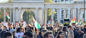A large crowd of people gathered in a public space, many holding flags and signs advocating for Palestine. The scene is framed by an architectural structure with pillars. The atmosphere is lively with a mix of ages and ethnicities present, all engaged in a demonstration.