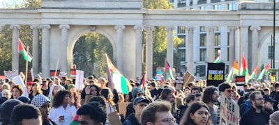 A large crowd of people gathered in a public space, many holding flags and signs advocating for Palestine. The scene is framed by an architectural structure with pillars. The atmosphere is lively with a mix of ages and ethnicities present, all engaged in a demonstration.