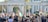 A large crowd of people gathered in a public space, many holding flags and signs advocating for Palestine. The scene is framed by an architectural structure with pillars. The atmosphere is lively with a mix of ages and ethnicities present, all engaged in a demonstration.