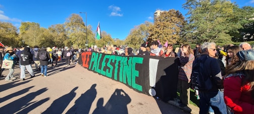 A group of people gather in an outdoor setting, holding a large banner with the text 'Free Palestine!' bolded in red, green, and white. The group stands on a path surrounded by trees, with some participants carrying backpacks and wearing jackets, indicating a cool day. Flags and signs are visible among the crowd.