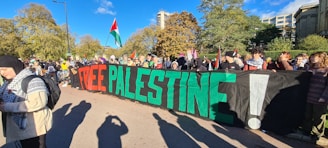 A large group of people gathered outdoors holding a banner with the words 'FREE PALESTINE' in vibrant red and green. Several individuals are holding or wearing Palestinian flags. The background features trees and tall buildings under a clear blue sky, suggesting an urban park setting. Attendees are diverse in appearance...