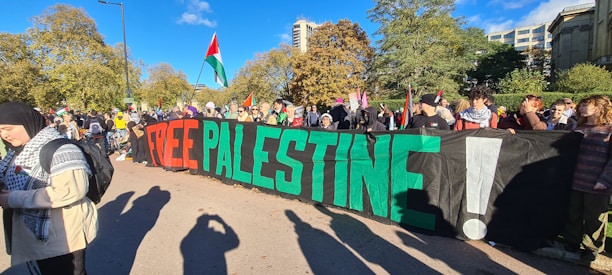 A large group of people gathered outdoors holding a banner with the words 'FREE PALESTINE' in vibrant red and green. Several individuals are holding or wearing Palestinian flags. The background features trees and tall buildings under a clear blue sky, suggesting an urban park setting. Attendees are diverse in appearance...