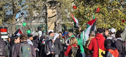 A group of people gathered outdoors holding flags and balloons. Many are wearing scarves, and some hold signs related to the Free Palestine movement. Trees and a decorative structure are visible in the background.