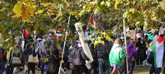 A group of people gathered outdoors, surrounded by trees with green and yellow leaves. Many participants are holding flags and signs, suggesting a protest or demonstration. The crowd is diverse, with individuals dressed in various colors, predominantly jackets, and hoodies.