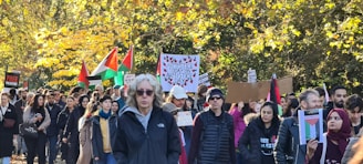 A large group of people are gathered outdoors, participating in a demonstration or protest. Many individuals are holding signs and banners with various messages, including flags with red, black, and green colors. The surrounding environment shows trees with autumn foliage, creating a colorful backdrop.