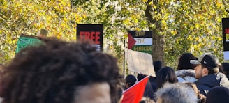 A crowd of people gathered outdoors in a wooded area, holding various protest signs with messages such as 'Free,' 'Stop the Genocide,' and others. The background is filled with green and yellow-leaved trees, and some people are wearing caps and hooded clothing.