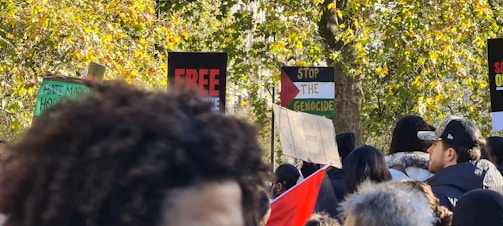 A crowd of people gathered outdoors in a wooded area, holding various protest signs with messages such as 'Free,' 'Stop the Genocide,' and others. The background is filled with green and yellow-leaved trees, and some people are wearing caps and hooded clothing.