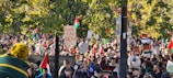 A large crowd gathers outdoors, holding signs and flags with various messages. The scene is filled with people, and many of the signs are related to political activism, such as one reading 'CEASE FIRE NOW.' The setting includes lush green trees, suggesting a park or garden area.