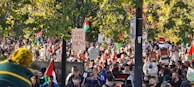 A large crowd gathers outdoors, holding signs and flags with various messages. The scene is filled with people, and many of the signs are related to political activism, such as one reading 'CEASE FIRE NOW.' The setting includes lush green trees, suggesting a park or garden area.
